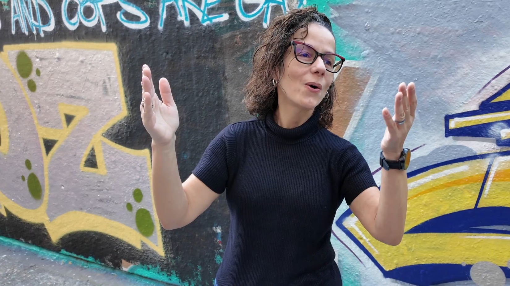 Aline Sardao mit erhobenen Händen vor einer farbigen Graffitiwand