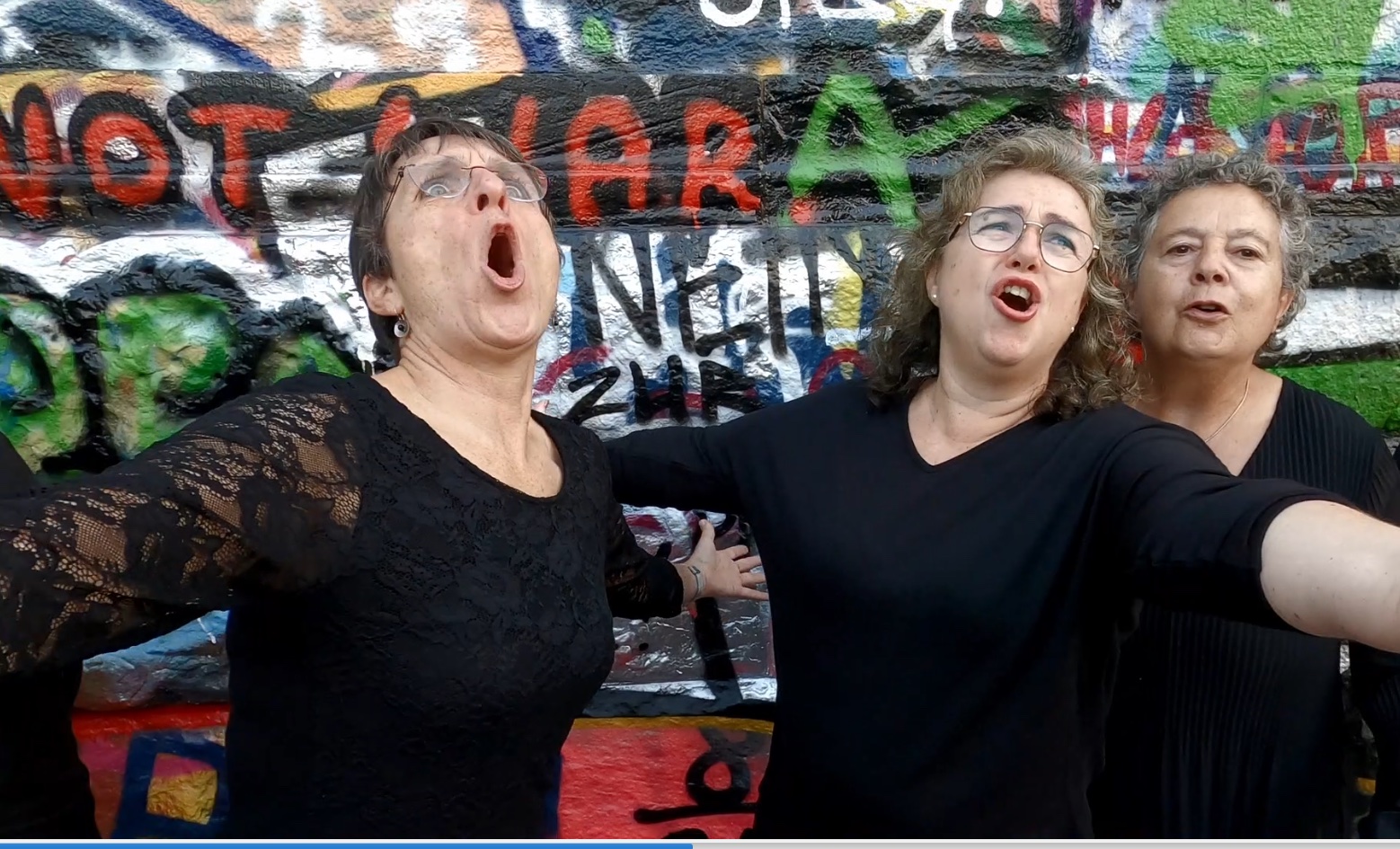 Drei Sängerinnen der RuhrChoriFeen singen mit ausgebreiteten Armen vor einer Graffitiwand