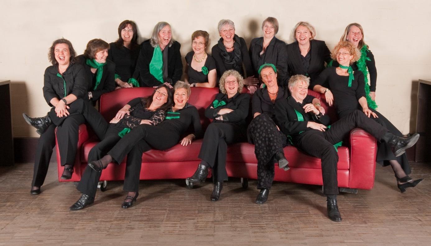Chorale Feminale in schwarzer Kleidung mit grünen Schals, gemeinsam auf und hinter einem roten Sofa